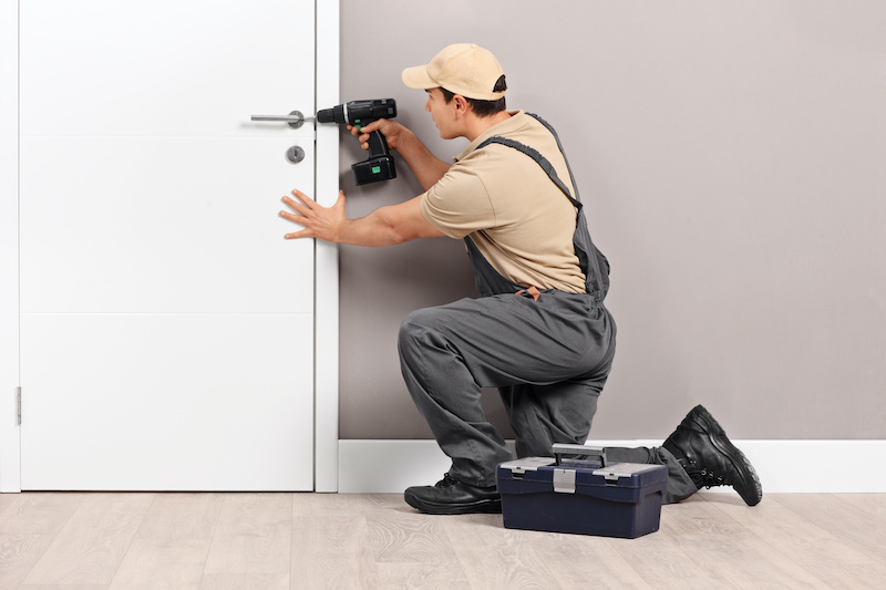 Young male locksmith installing a lock on a new white door with a hand drill